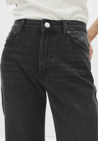 Personne portant un jean en denim noir taille haute et une chemise blanche, avec une main reposant sur sa hanche devant un fond uni.