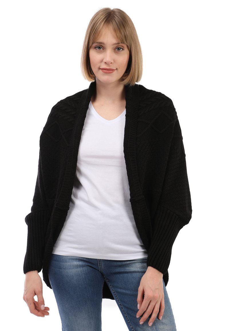 Cárdigan negro de punto con patrones texturizados, diseño oversize y puños acanalados. Lleva encima una camiseta blanca de cuello en V y jeans azules.