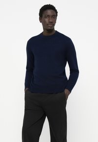 Navy blue knit sweater with a round neckline, long sleeves, and ribbed cuffs. Worn with black trousers, featuring a smooth texture.