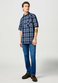 Camisa de cuadros azul de manga corta con detalles en blanco y marino, dos bolsillos en el pecho, combinada con jeans azules y zapatos marrones.