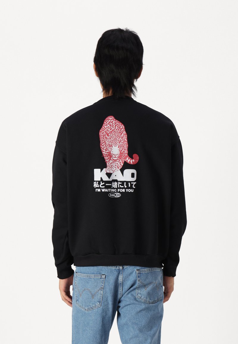 Svart hettegenser med et rødt leopardmotiv på baksiden, med hvit tekst som leser "Kao" og "JEG VENTER PÅ DEG" below.