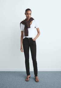 Jeans negros de cintura alta, diseño ajustado, combinados con una camiseta blanca de manga corta y un suéter de punto marrón caído sobre los hombros.