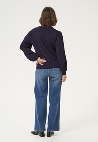 Marineblauer Pullover mit gerippten Puffärmeln, kombiniert mit hochtaillierten blauen Jeans und schwarzen Blockabsatzschuhen. Einfarbiger Hintergrund.