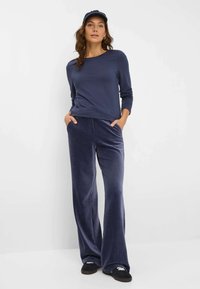 Marineblaues Langarmoberteil mit Rundhalsausschnitt, kombiniert mit weiten, weichen Velour-Hosen in Marineblau. Das Model trägt schwarze Schuhe und eine Marineblau-Kappe, die Hände in den Taschen.