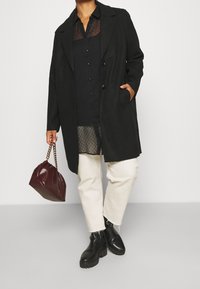 Persona che indossa un cappotto nero sopra una maglietta nera trasparente, pantaloni bianchi cropped, stivali neri, mentre tiene una borsa rosso scuro con tracolla in oro.