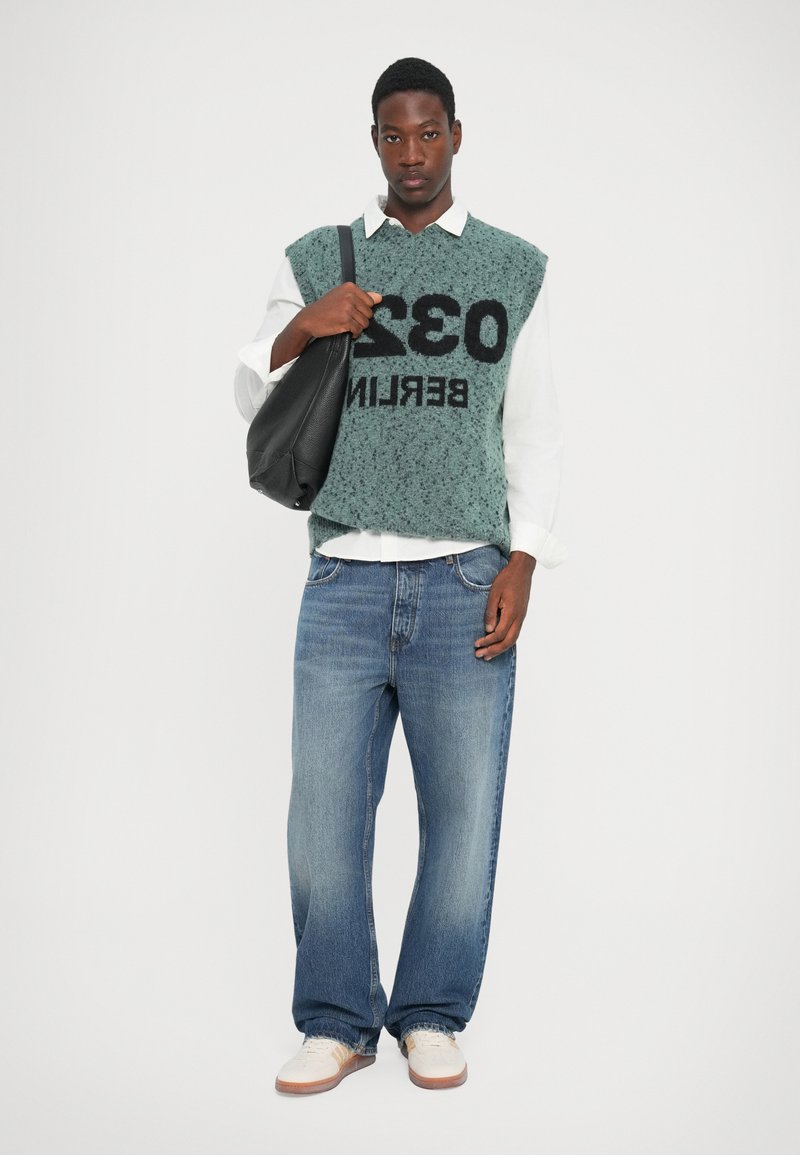 Khaki gebreide trui met bedrukt tekst, gecombineerd met blauwe losvallende jeans en witte sneakers. Een zwarte schoudertas completeert de outfit.
