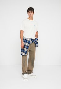 White cotton t-shirt with a small blue graphic, beige loose-fitting trousers, and a blue and white plaid shirt tied around the waist. White sneakers.
