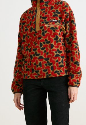 Persoon draagt een fleece pullover met rode, zwarte en tan patronen, voorzien van een borstzak met rits en tan drukknoopjes, gecombineerd met een zwarte broek.