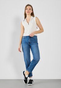 Hvit ermeløs zip-up topp med krage, paret med blå høytlivede jeans og svarte sneakers med hvite detaljer. Enkel, uformell antrekk.
