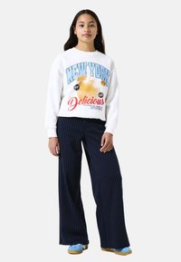 White sweatshirt with "NEW YORK Delicious" graphic, paired with navy striped wide-leg pants and light blue sneakers. Smooth fabric texture.