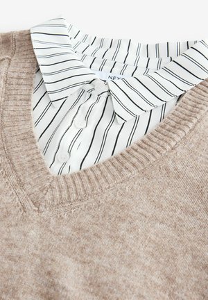 Beiger Strickpullover mit V-Ausschnitt. Darunter ein weißes Hemd mit Kragen, das schwarze vertikale Streifen und Knopfdetails aufweist.