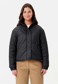 Doudoune noire avec un col montant, présentant des coutures diagonales et des poignets élastiques. Portée sur une chemise légère et associée à un pantalon beige.