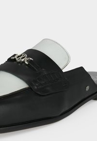 Chaussure noire en cuir à enfiler avec détail de boucle argentée et semelle intérieure blanche rembourrée, présentée sur un fond clair uni.