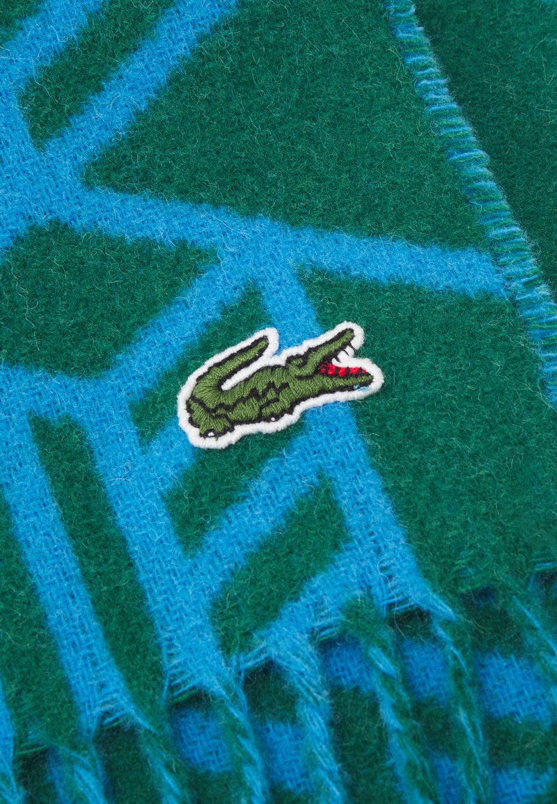 Bufanda de patrones verdes y azules hecha de material suave, con bordes con flecos y un logo de cocodrilo verde con detalles rojos.