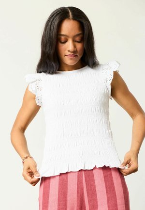 Frau mit schulterlangem dunklem Haar, die ein weißes, strukturiertes, ärmelloses Oberteil und rosa gestreifte Hosen trägt und den Saum des Oberteils richtet.