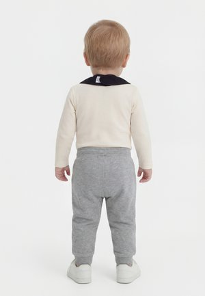 Un tout-petit est debout de dos, portant un haut à manches longues crème avec un col noir, un pantalon gris et des baskets blanches sur un fond blanc.