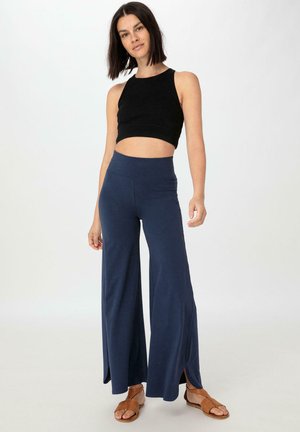 Schwarzes, kurzes Tanktop kombiniert mit marineblauen, weit geschnittenen Hosen mit seitlichen Schlitzen. Strukturierter Stoff mit glattem, atmungsaktivem Design und braunen Sandalen.
