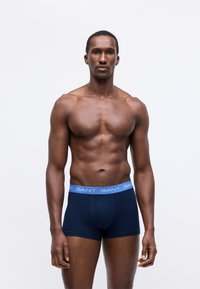 Uomo adulto con un fisico tonico che indossa boxer blu navy con una cintura azzurra chiara etichettata "GANT", in piedi su uno sfondo bianco.