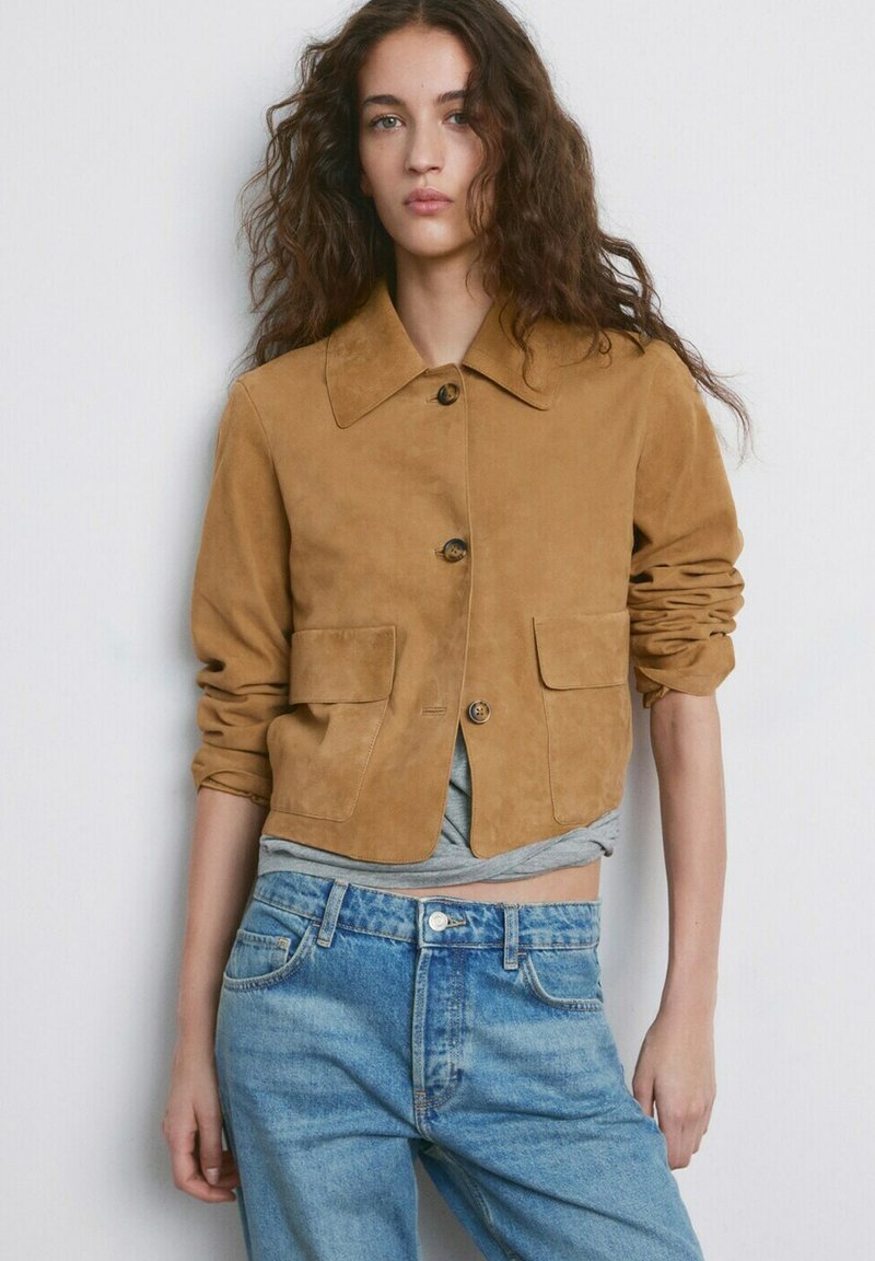 Mladá žena s dlouhými kudrnatými vlasy nosí světle hnědou semišovou bundu na knoflíky a světle modré džíny s vysokým pasem, stojí na bílém pozadí.