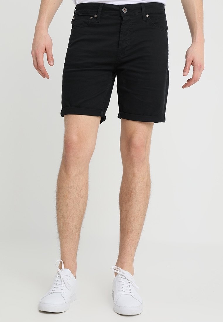 Homme portant un short noir à ourlet roulé et des baskets blanches, se tenant devant un fond clair uni.