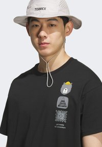 T-shirt noir avec un graphique d'ours portant un chapeau jaune et du texte. Chapeau seau blanc avec le logo "TERREX". Les deux sont des vêtements décontractés pour activités de plein air.