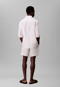 Mann trägt ein weißes Langarmshirt mit hochgekrempelten Ärmeln, weiße Shorts und schwarze Slip-On-Sandalen, steht mit dem Rücken zur Kamera vor einem grauen Hintergrund.