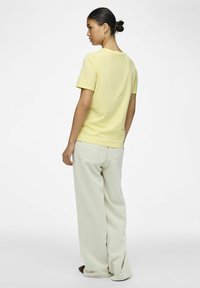 Tricou galben din bumbac cu mâneci scurte și guler clasic rotund, asortat cu pantaloni bej deschis, largi, cu model în relief, văzuți din spate.