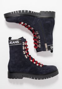 Námořnické nubukové kotníkové boty s červenými tkaničkami, stříbrnými kovovými očky, bočním zipem a robustní černou podrážkou s hlubokými vzory. Název "JEANS" na vrchu.
