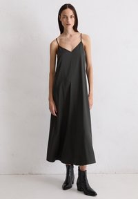 Langes, dunkelgrünes Slipkleid mit dünnen Trägern, V-Ausschnitt und fließender Silhouette, kombiniert mit schwarzen Knöchelstiefeln. Glatte Textur.