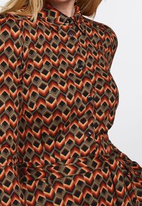 Camicia abbottonata con un motivo geometrico in tonalità di rosso, arancione e nero, dotata di colletto e bottoni neri sulla parte frontale.