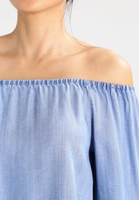 Top azzurro chiaro con spalle scoperte realizzato in tessuto morbido, con scollatura elastica arricciata e maniche fluide, caratterizzato da una texture liscia.