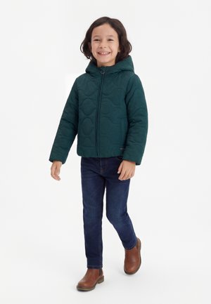 Niño sonriente con cabello hasta los hombros caminando, vestido con una chaqueta acolchada con capucha de color verde oscuro, vaqueros oscuros y botines marrones.