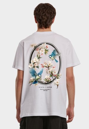 Person som har på seg hvit T-skjorte med blomstermotiv og to blå fugler inni en oval ramme, med teksten "Kyoto / Japan" under.