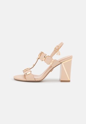 Beige korkokengät, joissa on geometrian muotoinen tukikorko, säädettävä nilkkavyö ja koristeelliset metalliset yksityiskohdat. Sujuva nahkatuntuma.