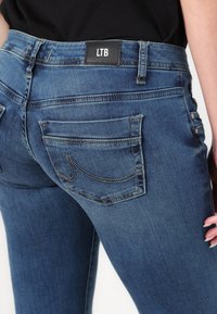 Jeans de mezclilla azul con un diseño ajustado. Las características incluyen dos bolsillos traseros y acentos cosidos. Logo de LTB en un parche de cuero negro en la cintura.