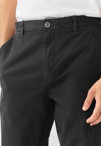 Close-up van een persoon die zwarte knoopsluiting broek en een witte ingeklede shirt draagt, met één hand rustend nabij de bovenbeen.