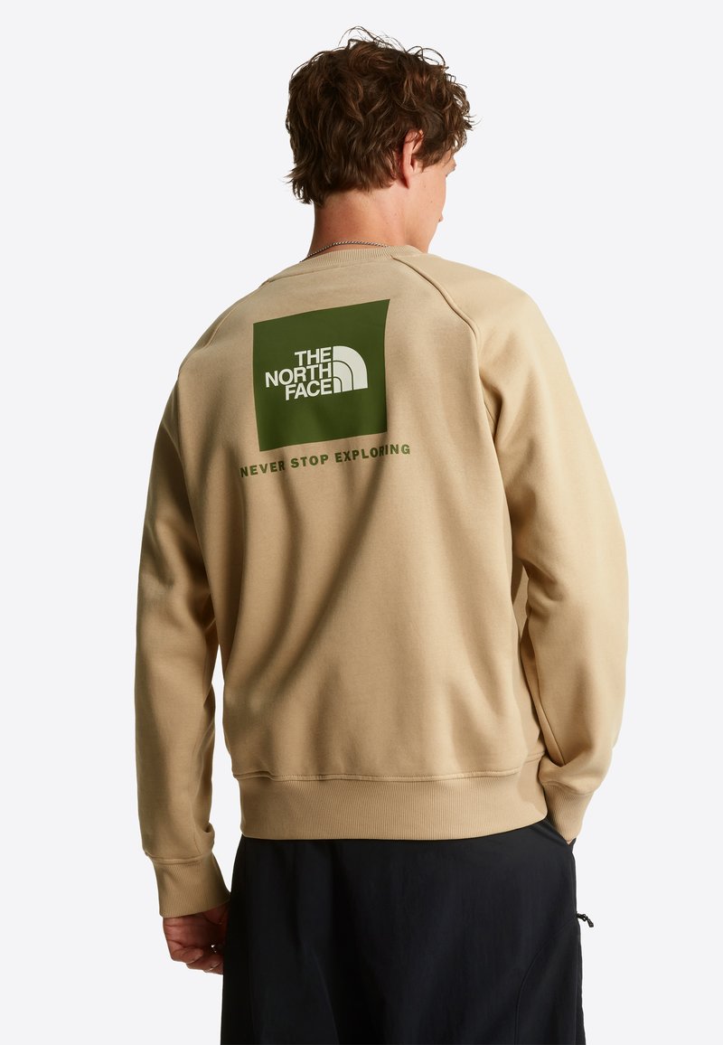 Man wearing beige sweatshirt with green The North Face logo and "Never Stop Exploring" text on the back, paired with black pants.