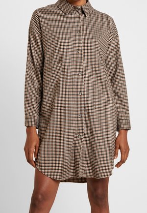 Sukienka koszulowa z długim rękawem i zapięciem na guziki z przodu, w brązowo-czarno-białą kratę, noszona przez osobę stojącą nieruchomo.