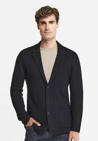 Mörkblå stickad kavaj med sjalkrage, tre knappar och två framfickor, bärs över en ljusbeige skjorta. Slät textur och skräddarsydd passform.