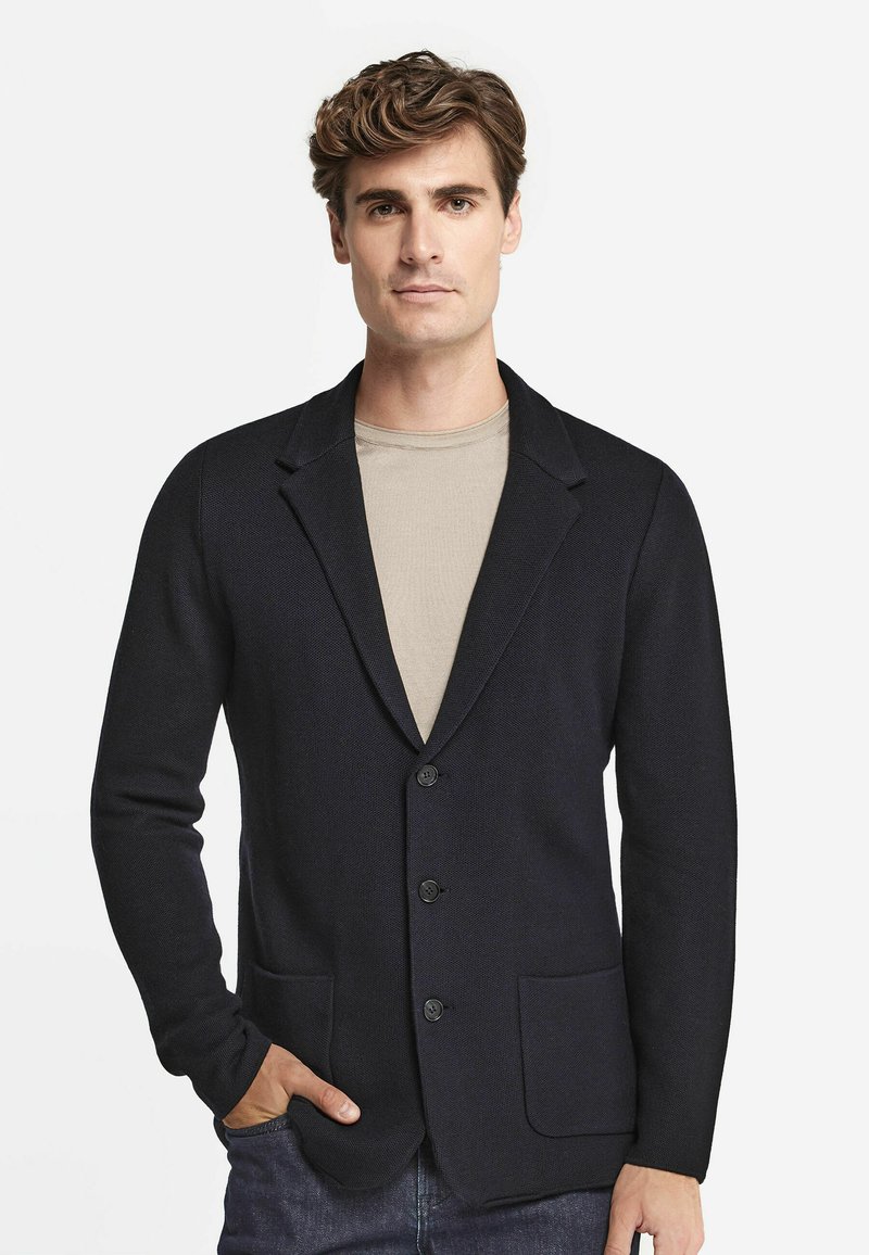 Mörkblå stickad kavaj med sjalkrage, tre knappar och två framfickor, bärs över en ljusbeige skjorta. Slät textur och skräddarsydd passform.