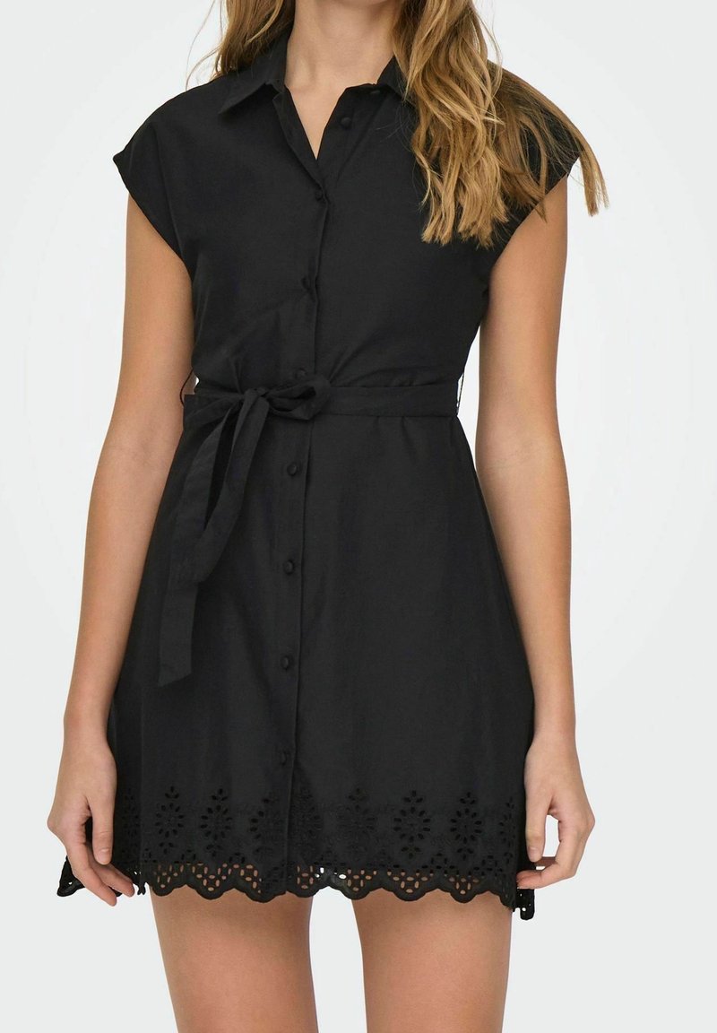 Robe noire à manches courtes boutonnée avec ceinture à la taille et ourlet festonné à découpe florale portée par une personne debout aux longs cheveux.