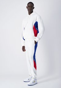 Un sweat à capuche blanc et un pantalon avec des panneaux latéraux bleus et rouges. Le sweat a un cordon de serrage ; le pantalon possède des poches. Portés avec des chaussures blanches.