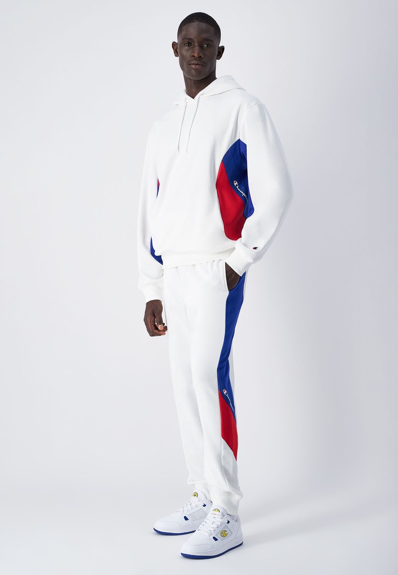 Felpa bianca con cappuccio e pantaloni con pannelli laterali blu e rossi. La felpa ha un cordoncino; i pantaloni presentano tasche. Indossati con scarpe bianche.