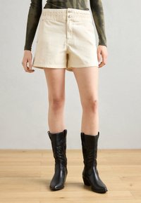 Beige cotton shorts with a braided waistband, front buttons, and two side pockets, worn with black leather cowboy boots.
