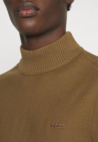 Gros plan sur une personne portant un pull marron côtelé à col cheminée avec « BOSS » brodé sur la poitrine.