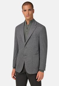 Šedý houndstooth blazer s klopami, zapínáním na dva knoflíky, našitými kapsami a texturovanou tkaninou. Nesený přes zelený zipový top.