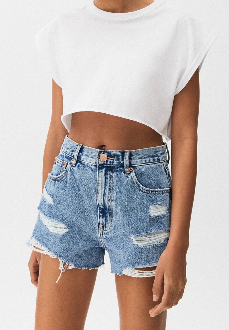Personne portant un débardeur court blanc et un short en jean bleu taille haute avec des détails usés et des ourlets effilochés.