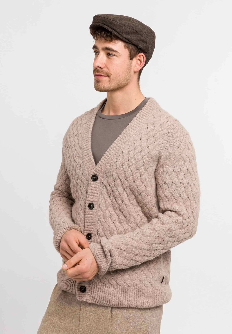 Man in een bruine flat cap, met een beige kabelgebreide cardigan met knopen over een bruine shirt, staand met de handen in elkaar tegen een witte achtergrond.