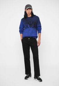 Fiatal férfi kék Versace Jeans Couture pulóverben, fekete nadrágban fehér varrásokkal, fekete-fehér sportcipőben és fekete sapkában.