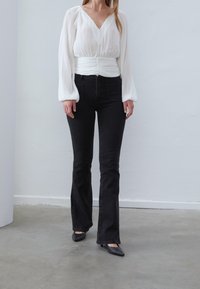 Blusa blanca de manga larga con pliegues, escote en V y cintura fruncida, combinada con jeans negros de campana y zapatos negros de punta.